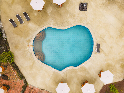 Aerial view of a turquoise swimming pool surrounded by lounge chairs and white umbrellas on a paved patio in Santa Cruz, Galapagos Province, Ecuador.