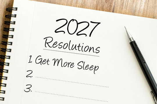 Handwritten 2027 resolutions list including get more sleep on a notebook page next to a black pen on a wooden surface.
