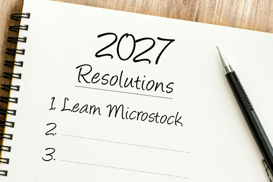 Handwritten 2027 resolutions list including learn microstock on a notebook page next to a black pen on a wooden surface.