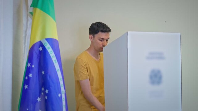 Jovem eleitor vota na cabine eleitoral, com a bandeira do Brasil desfocada em primeiro plano, durante as elei&ccedil;&otilde;es presidenciais de outubro.
