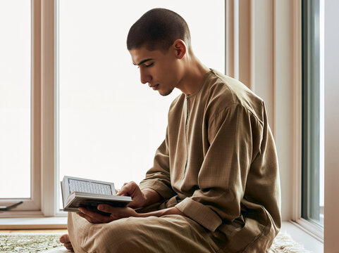 Muslim man reading the Quran in a bright home prayer area. Young Middle Eastern man in thobe sitting on a rug studying the holy book during Ramadan