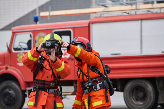 Two Firefighters In Protective Gear Using Thermal Camera In Front Of Red Fire Truck During Rescue