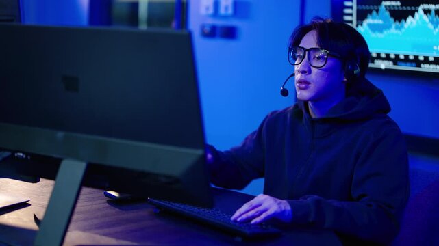 Focused Asian male day trader in a hoodie and headset working late at night in a dark blue lit room. He is talking and typing on his computer, analyzing financial crypto and stock market charts.