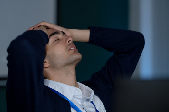 Adult male technology professional holds head with eyes closed beside computer monitor in modern office, expressing stress from software troubleshooting, cybersecurity review, and digital workload.