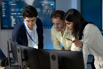 Three adult technology professionals lean toward monitors in modern office, reviewing software system together while female colleague points during cybersecurity analysis and problem solving. © Ekkasit A Siam