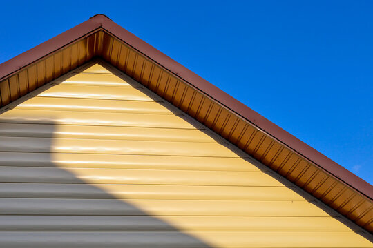 The gable of a modern house is finished with beige vinyl siding and brown soffit. The gabled roof features crisp geometric lines against a clear blue sky. Bright sunlight casts a distinct sharp shadow