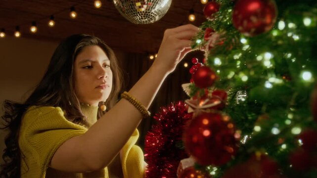 Hispanic young woman decorating Christmas tree with festive hanging ornaments and baubles. Clip ideal for winter holiday advertising, seasonal promos, and Christmas family lifestyle commercials