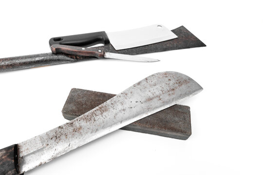 Old rusty machete knife signs of wear from heavy use resting on whetstone sharpening stone and set various knives, meat cleaver isolated on white background. tools maintenance concepts.