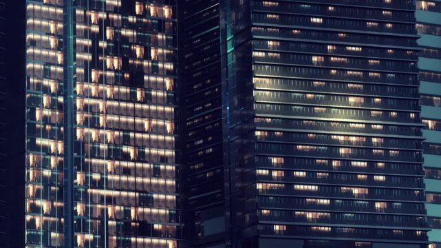 Tall buildings stand side by side in a city. Many windows are lit up at night. The scene shows a busy urban environment with various structures.