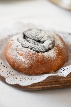 Fresh baked poppy seed bun dusted with powdered sugar, served on wooden plate with lace napkin in cozy minimalist setting.