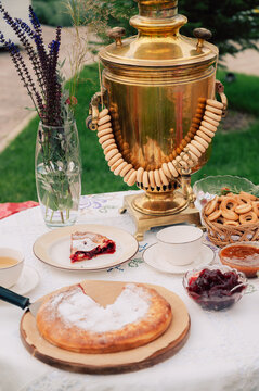A golden samovar with bagels, tea cups, homemade pie, and jam on a white tablecloth set for tea in a garden.