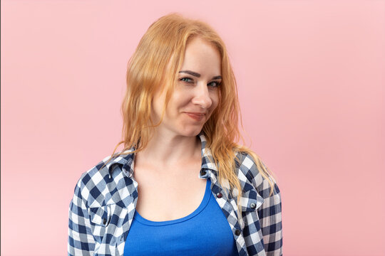 Young woman with red hair and a checkered shirt is playfully smirking at the camera, expressing confidence, allure, and a mischievous attitude against a pink background