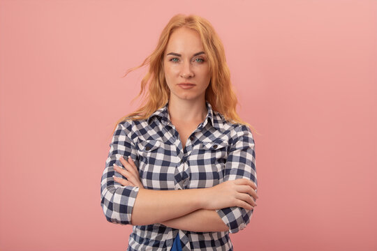 Young woman standing confidently with a serious facial expression and arms crossed, conveying strength, determination, and perhaps an air of disapproval or skepticism against a solid pink background