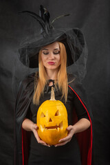 Young woman in witch hat and black cape holding a traditional carved jack o' lantern, celebrating the autumn spooky holiday season © somemeans