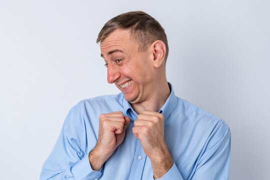 Man feeling shy and awkward, clutching his shirt collar and fists to his chest while smiling sheepishly, displaying playful embarrassment and joyful, comical expression on white background