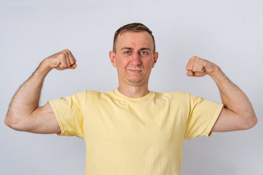 Man in a yellow t shirt standing against a white background, flexing his arm muscles, showing physical strength, determination, and a sense of accomplishment