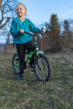 Young blonde girl with a green and black bike stands on a grassy hillside, gazing ahead and ready for outdoor play and learning to ride, joyful and adventurous