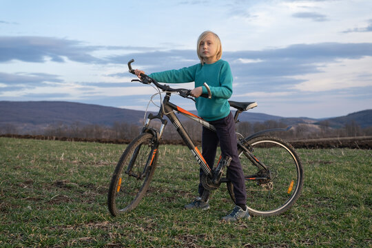 Young boy with blonde hair standing next to a black and orange bicycle in a green grass field, looking directly at the viewer with hills and a cloudy sky in the background