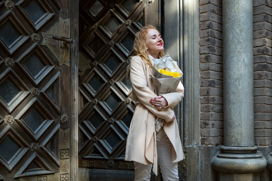 Woman standing in warm sunlight embracing joy and appreciation, holding a bouquet of bright yellow flowers in front of an ornate door