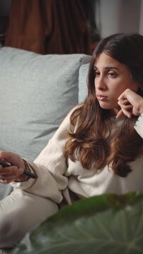 Young Latina woman sitting bored on her sofa while watching television, representing entertainment, leisure, media consumption, and everyday home lifestyle.
