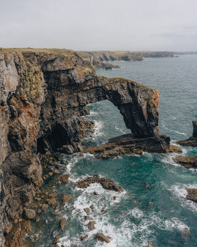 View of the Green Bridge of Wales, a natural limestone arch formation rising above the turquoise sea with rugged coastal cliffs and crashing waves, Pembroke, United Kingdom.