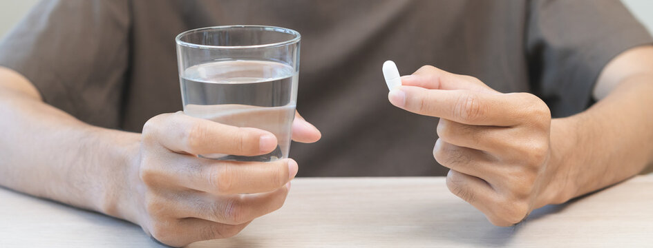 Sick asian young man holding tablet pill on hand, painkiller medicine from fever, stomach pain, headache for treatment, take drug, vitamin with glass of water on table at home. Health care concept.