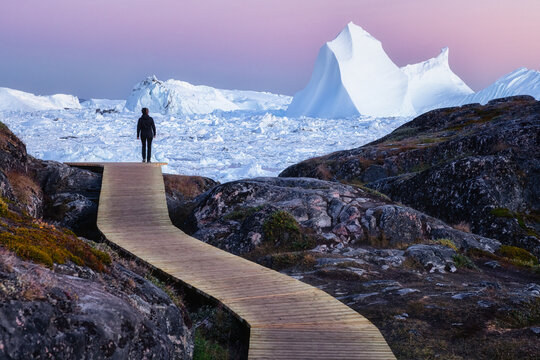 Person standing on a wooden path looking at giant icebergs with pink sky above