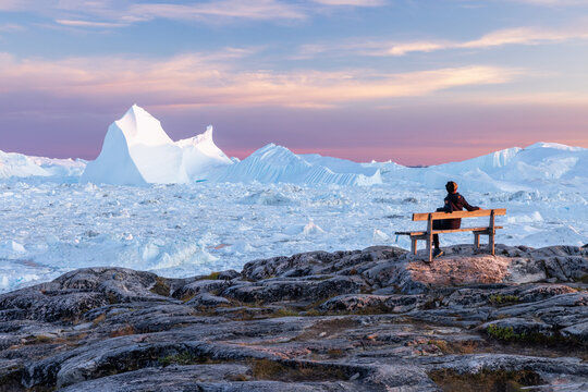 Person sitting on a wooden bench with views in nature looking at giant icebergs in Ilulissat icefjord in Greenland