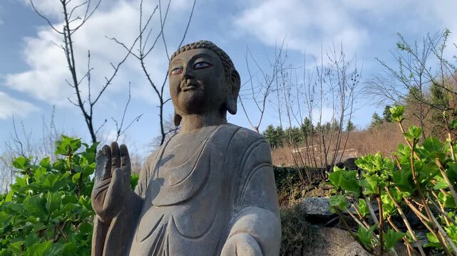 A stone Buddha statue in nature. In the background are plants and a blue spring sky.