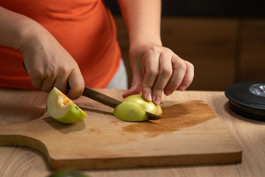 Woman slices a fresh green apple on a wooden cutting board in the kitchen, with her hands holding the fruit and knife.