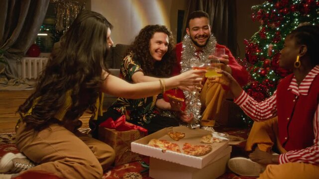 Multiethnic young adults toasting drinks and eating pizza while sitting near Christmas tree during holiday party. Footage suitable for food delivery and winter holiday promotions