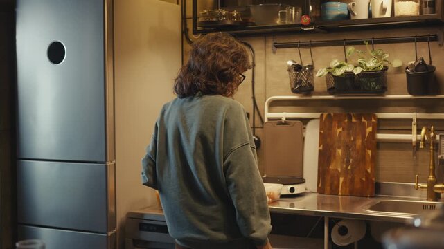 Young woman from behind entering dark loft kitchen at night and turning on under-cabinet LED lighting to illuminate metal countertop with sink and portable induction stove before cooking dinner.