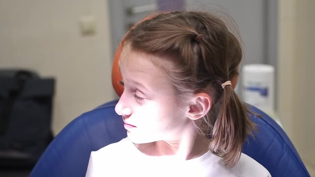 Anxious young girl sits in dental chair during checkup, looking down with tense expression under bright examination light.