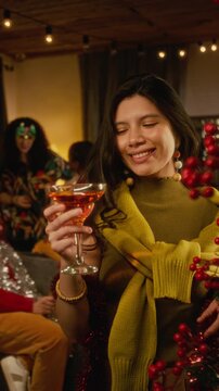 Vertical portrait of smiling young Hispanic woman toasting with cocktail glass during holiday celebration. Ideal for seasonal beverage commercials and festive event marketing campaigns