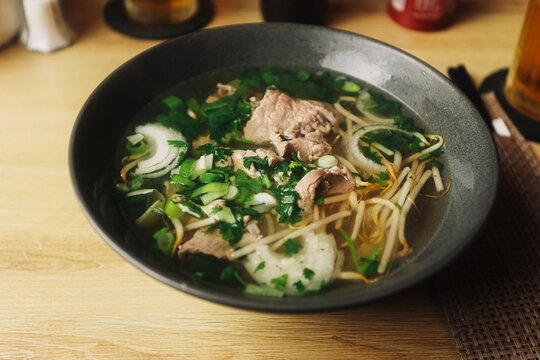 Traditional Asian noodle soup with sliced beef bean sprouts onion and fresh herbs served in a dark bowl. Hot clear broth and noodles create a comforting restaurant food background.
