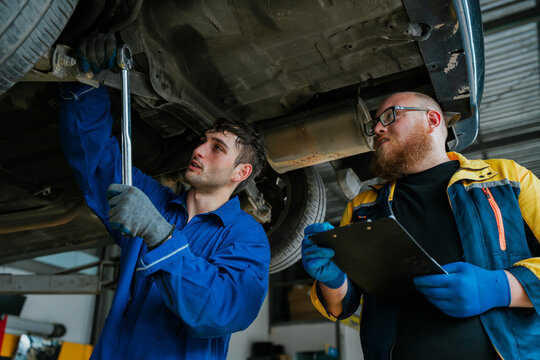 Focused car mechanic performing auto repair in garage shop with happy colleague checking clipboard under vehicle maintenance service
