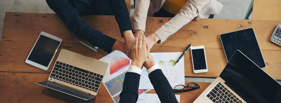 Group of businesswoman joining and hold in hands during business meeting at wooden table in indoor office, surrounded by laptops, tablets, documents and charts, showing teamwork, cooperation and unity