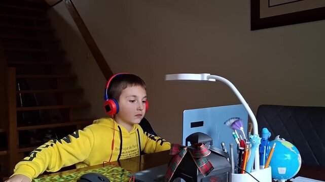 7 year old boy at home using a computer with headphones on a desk with school supplies