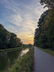 Fototapeta premium Tranquil Canal Towpath at Sunset with Tree-Lined Waterway and Dramatic Golden Sky