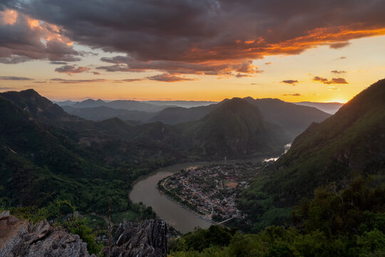 Aerial view of the village of Nong Khiaw nestled along the Nam Ou River, surrounded by limestone karst mountains under a vibrant sunset sky, Nong Khiaw, Luang Prabang Province, Laos.