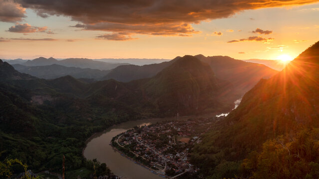 Aerial view of Nong Khiaw nestled along the Nam Ou River surrounded by lush mountains at sunset with golden light reflecting on the water in Nong Khiaw, Luang Prabang Province, Laos.