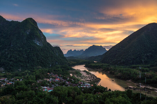 Aerial view of the village of Nong Khiaw nestled between lush green karst mountains along the Nam Ou River during a vibrant sunset in Nong Khiaw, Luang Prabang Province, Laos.