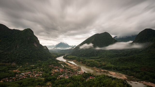 Aerial view of the Nam Ou river winding through the lush green mountains and the village of Nong Khiaw under a cloudy sky in Nong Khiaw, Luang Prabang Province, Laos.