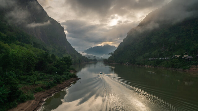 Aerial view of a boat traveling along the Nam Ou river surrounded by lush green mountains and dramatic clouds in Nong Khiaw, Luang Prabang Province, Laos.