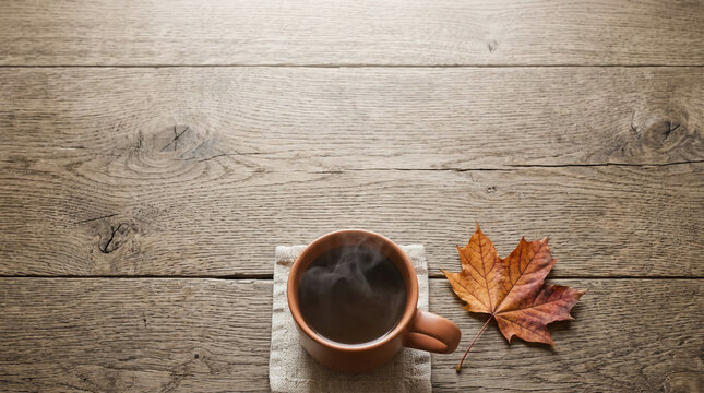 Steaming ceramic mug with hot coffee on rustic wooden surface beside a fallen autumn leaf. Warm natural light illuminates a cozy seasonal scene for a peaceful morning beverage break.