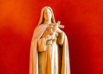 Naklejka premium A white statue of Saint Thérèse holds a crucifix with roses before a red wall.