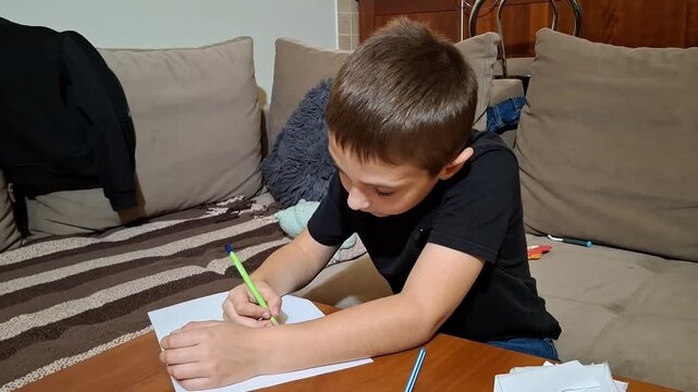 Seven year old boy draws at home while sitting at a table with papers and pens around him