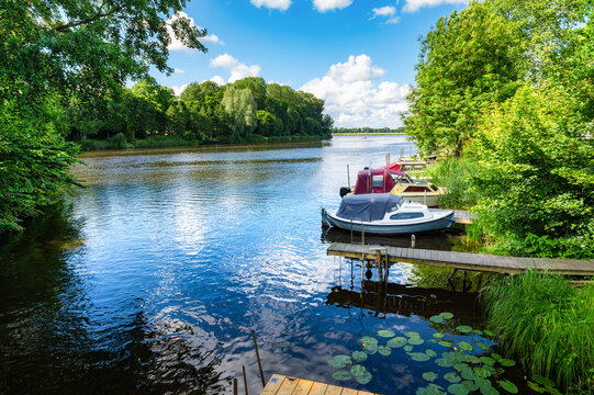 Small recreational boats are moored at wooden jetties near Friedrichstadt, surrounded by trees, reeds, water lilies and lush summer greenery.