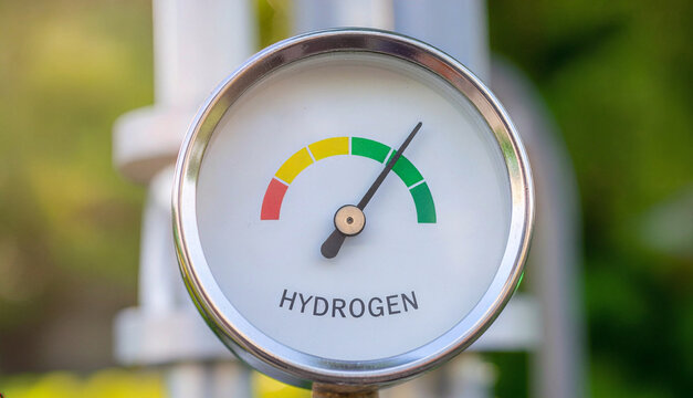 A close up of a round pressure gauge with hydrogen text showing a measurement needle pointing to the green zone against a blurred outdoor background, representing clean energy control