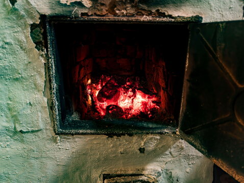 Rustic fireplace with glowing red embers providing warm, ambient light. The hearth shows signs of use with soot-stained bricks and surrounding textured, weathered plaster walls. Metal door slightly aj